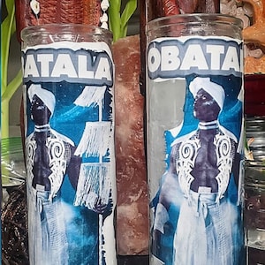 Può includere: Due portacandele di vetro con immagini blu e bianche di figure che tengono bastoni. Le figure indossano abiti bianchi e hanno la pelle scura. I testi "OBATALA" e "ATALA" sono stampati sulla parte anteriore dei portacandele. Una pietra rotonda bianca è in primo piano.