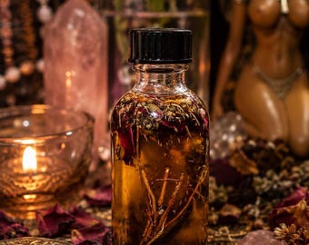 May include: A clear glass bottle with a black screw-top lid, filled with a golden-brown liquid containing visible dried botanical elements, including small twigs, dark herbs, and scattered flower petals suspended throughout the oil.