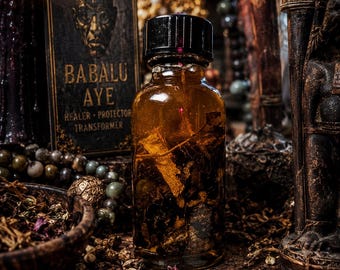 May include: A clear glass bottle with a black cap containing amber liquid and botanical matter. Behind it stands a rectangular card featuring a portrait of a figure with the text BABALU AYE HEALER PROTECTOR TRANSFORMER in gold lettering.