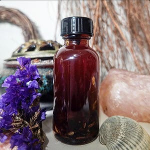 May include: A dark red glass bottle with a black cap, filled with a dark liquid and small particles. The bottle is surrounded by purple flowers, a seashell, and a pink crystal. The background includes a decorative object and dried plants.