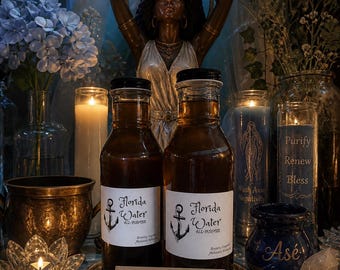 May include: Two bottles of Florida Water All-Purpose with anchor labels sit in front of a statue. A card reads "Purification: Peace, Clarity, Renewal." The scene includes candles, flowers, and a blue jar with "Asé" on it.