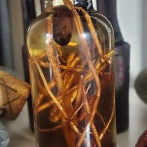 Könnte beinhalten: Eine klare Glasflasche mit schwarzem Deckel, gefüllt mit einer goldenen Flüssigkeit und braunen Wurzeln. Die Flasche steht auf einer weißen Oberfläche.