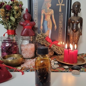May include: A collection of spiritual items including a framed Egyptian art print, a wooden statue, candles, dried flowers, and a glass bottle filled with liquid. Other items include jars of pink salt and rose petals, and a small brass bowl.