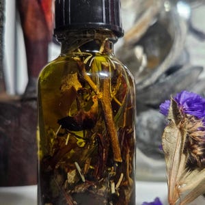 Puede incluir: Una pequeña botella de vidrio llena de un líquido transparente y varias hierbas y materiales vegetales. La botella tiene una tapa negra y está sobre una superficie blanca.