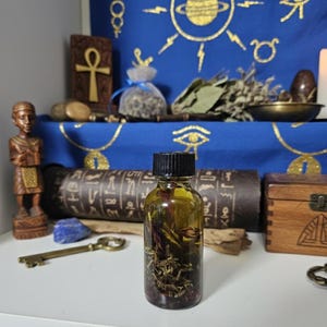 Puede incluir: Una botella de vidrio transparente llena de líquido dorado y hierbas, con tapa negra. Se encuentra en un estante blanco con una caja de madera, una llave y un fondo azul. Otros objetos espirituales.