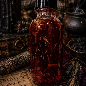 May include: A clear glass bottle with a black cap filled with a dark red liquid and botanical inclusions. Beside the bottle lies a rectangular parchment card printed with the text BAD LUCK EVIL EYE CURSES UNDO & REVERSE and an eye icon.