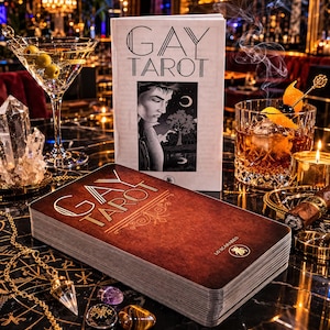 May include: A deck of "GAY TAROT" cards with a brown cover and gold lettering, next to a matching book. A martini with olives and an old fashioned cocktail are also in the image, along with crystals, a cigar, and a lit candle.