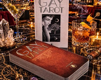 May include: A deck of "GAY TAROT" cards with a brown cover and gold lettering, next to a matching book. A martini with olives and an old fashioned cocktail are also in the image, along with crystals, a cigar, and a lit candle.