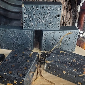 May include: Four black bars of soap with intricate gold designs. The designs include a butterfly, a sun, and a crescent moon. The soap bars are wrapped in sheer black fabric with gold stars.