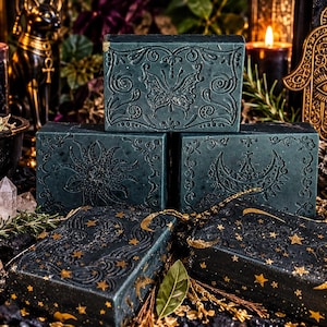 May include: Five rectangular dark green soap bars featuring embossed designs of a butterfly, a sun, a crescent moon, and gold-painted celestial patterns including stars and moons, arranged in a stack on a dark surface with various decorative items.