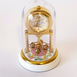 May include: A gold-toned clock with a clear glass dome. The clock face features an illustration of two figures. The base is white with a floral design. The clock is approximately 5 inches tall.