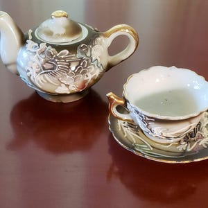 May include: A miniature tea set featuring a teapot and teacup with saucer. The set is primarily cream and dark green with gold accents. The teapot and teacup have a detailed relief design. The teacup sits on a matching saucer.