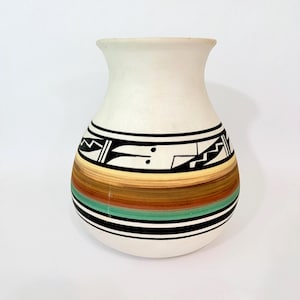 May include: A white ceramic vase with a wide base and a narrower neck. The vase features a band of geometric black and white designs above a series of horizontal stripes in shades of brown, green, and orange. The vase is approximately 8 inches tall.