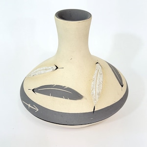 May include: Cream-colored ceramic vase with a gray rim and base. The vase features a band of gray with feather designs and an arrow. The vase has a rounded body and a narrow neck.