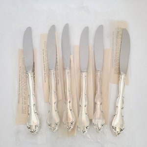 May include: Six silver butter knives in individual plastic packaging. The knives have ornate handles and are packaged with instructions for care.