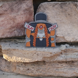 May include: A black and orange iron-on patch featuring a cartoon illustration of a woman wearing a black hat and a black dress. The woman is holding up two signs that say "I SLAY" and "OKAY".