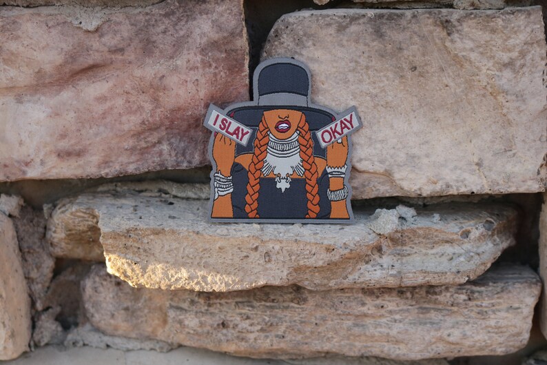 May include: A black and orange iron-on patch featuring a cartoon illustration of a woman wearing a black hat and a black dress. The woman is holding up two signs that say "I SLAY" and "OKAY".
