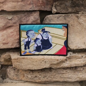 May include: Embroidered patch featuring a cartoon scene with three characters in blue and black outfits, set against a light blue background. The patch has a black border and is displayed against a textured stone wall.