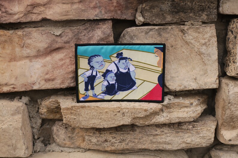 May include: Embroidered patch featuring a cartoon scene with three characters in blue and black outfits, set against a light blue background. The patch has a black border and is displayed against a textured stone wall.