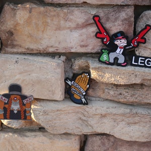 May include: Three embroidered patches on a stone wall. The first patch features a cartoon character holding two guns and a bag of money with the word "LEGACY" below. The second patch features a pair of hands in a praying position with the text "SOCIALLY AWKWARD". The third patch features a cartoon character with a hat and the text "I SLAY OKAY".
