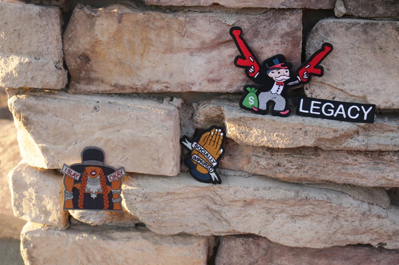 May include: Three embroidered patches on a stone wall. The first patch features a cartoon character holding two guns and a bag of money with the word "LEGACY" below. The second patch features a pair of hands in a praying position with the text "SOCIALLY AWKWARD". The third patch features a cartoon character with a hat and the text "I SLAY OKAY".