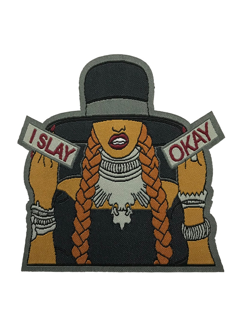 May include: Embroidered iron-on patch featuring a cartoon illustration of a woman with long braids wearing a black hat and a black dress. The woman is holding two signs that say "I SLAY" and "OKAY".