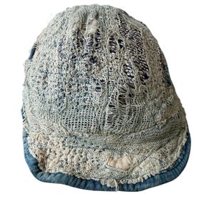 May include: A distressed, light blue denim cap with a unique, open-weave design. The cap features a frayed, textured appearance with visible holes and a contrasting dark blue brim. This vintage-style hat offers a worn, artistic aesthetic.