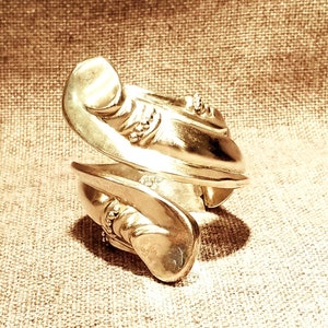 May include: A silver-toned, twisted metal bangle bracelet with decorative details. The bracelet is on a textured, beige fabric background.