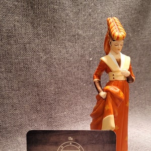 May include: A porcelain figurine of a woman wearing a red and yellow dress and a matching headpiece. The figurine is standing on a wooden surface with a business card that reads "Its Vintage Sage" and a website address.