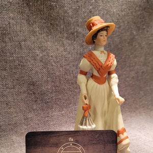 May include: A porcelain figurine of a woman wearing a white dress with orange accents and a straw hat. She is holding a white parasol with a red bow. The figurine is standing on a wooden surface with a business card that reads "Its Vintage Sage" and a website address.