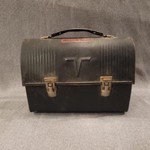 Könnte beinhalten: Eine schwarze Vintage-Metall-Lunchbox mit Griff und zwei Verschlüssen. Die Lunchbox hat ein geriffeltes Design auf der Oberseite und ein großes "T"-Emblem auf der Vorderseite. Ein kleines rotes Etikett ist oben sichtbar. Die Lunchbox stammt wahrscheinlich aus der Mitte des 20. Jahrhunderts.
