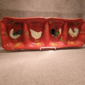 Puede incluir: Un plato de servir de cerámica roja con cuatro secciones, cada una con un diseño de gallo pintado a mano. Los gallos son blancos, negros y verdes con acentos amarillos. El plato tiene un borde festoneado y es perfecto para servir aperitivos o bocadillos.