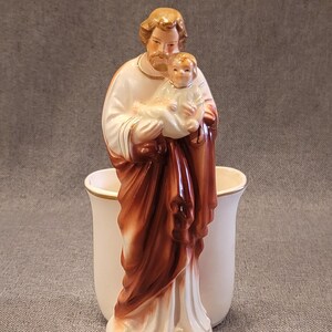 May include: A ceramic figurine of Saint Joseph holding the baby Jesus. The statue is primarily white, with brown and gold accents. The figurine is placed in front of a white vase with a gold rim.