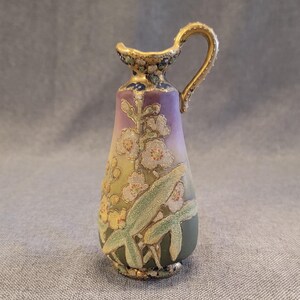 May include: A small, gold-trimmed porcelain pitcher with a floral design. The pitcher has a light purple, yellow, and green gradient with white flowers and green leaves.