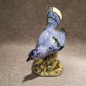 May include: A blue and white ceramic figurine of a blue jay perched on a branch. The bird has a white crest and black markings on its wings.