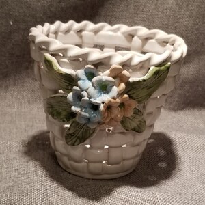 May include: A white ceramic basket-style planter with a woven design. The planter is adorned with a cluster of blue, tan, and green floral accents. The basket has a round shape and is suitable for small plants or decorative use.