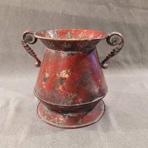 Könnte beinhalten: Eine rot-verwitterte Metallvase mit zwei verzierten Griffen. Die Vase hat eine breite Öffnung und einen dekorativen Sockel. Goldakzente und Fleur-de-Lis-Muster zieren den Rand und verleihen diesem Einrichtungsgegenstand im Vintage-Stil einen Hauch von Eleganz.