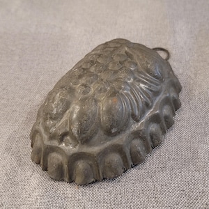 May include: A vintage, oval-shaped metal mold with a detailed floral design on the top surface. The mold is a dark gray color and has a small loop for hanging. The edges have a scalloped design.