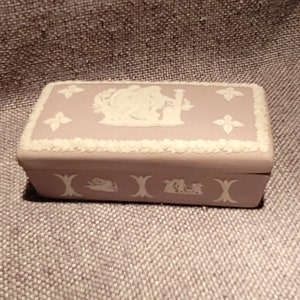 May include: A lavender colored ceramic trinket box with white raised designs. The lid features a scene of two figures in a garden setting. The bottom of the box has a repeating design of stylized trees and figures.