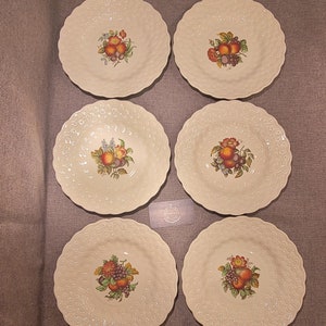 May include: Six off-white ceramic plates with a scalloped edge and a raised dot pattern. Each plate features a central fruit design with peaches, grapes, and other fruits in shades of red, orange, and purple.