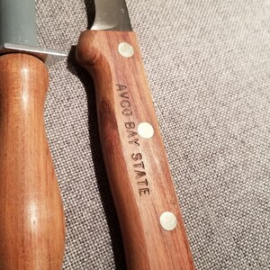 May include: A wooden handled knife with a stainless steel blade. The handle is engraved with the words "AVCO BAY STATE".