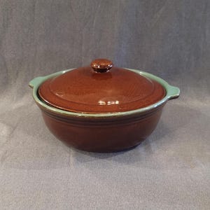 May include: A brown ceramic casserole dish with a matching lid. The dish has a light blue rim and handles. The lid has a small knob. The dish is likely used for cooking and serving food.