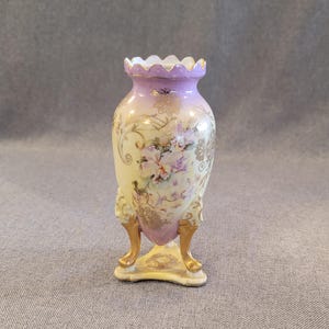 May include: A small, ornate vase with a scalloped rim and three gold-colored feet. The vase has a light yellow base with a lavender top and gold accents. Floral and gold leaf designs adorn the body of the vase, creating an antique aesthetic.