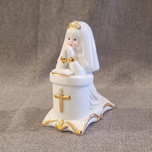 May include: White porcelain figurine of a girl kneeling in prayer at an altar with a gold cross. She is wearing a white veil and a gold crown.