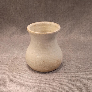 May include: A small, off-white ceramic vase with a rounded body and a slightly flared rim. The vase has a speckled texture and a smooth, glazed finish. The neutral color and simple design make it suitable for various decor styles.