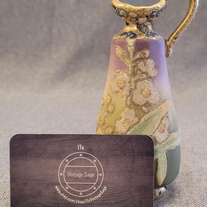May include: A small, hand-painted porcelain pitcher with a floral design in shades of purple, green, and gold. The pitcher has a handle and a spout, and it stands on three small feet.