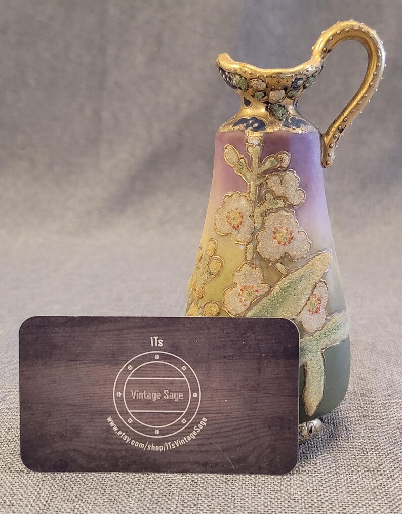 May include: A small, hand-painted porcelain pitcher with a floral design in shades of purple, green, and gold. The pitcher has a handle and a spout, and it stands on three small feet.