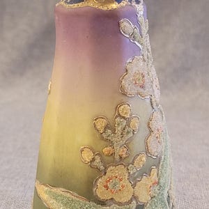 May include: A small, hand-painted ceramic pitcher with a gold handle and floral design. The pitcher has a gradient of colors from purple to yellow to green, with gold accents and white flowers.