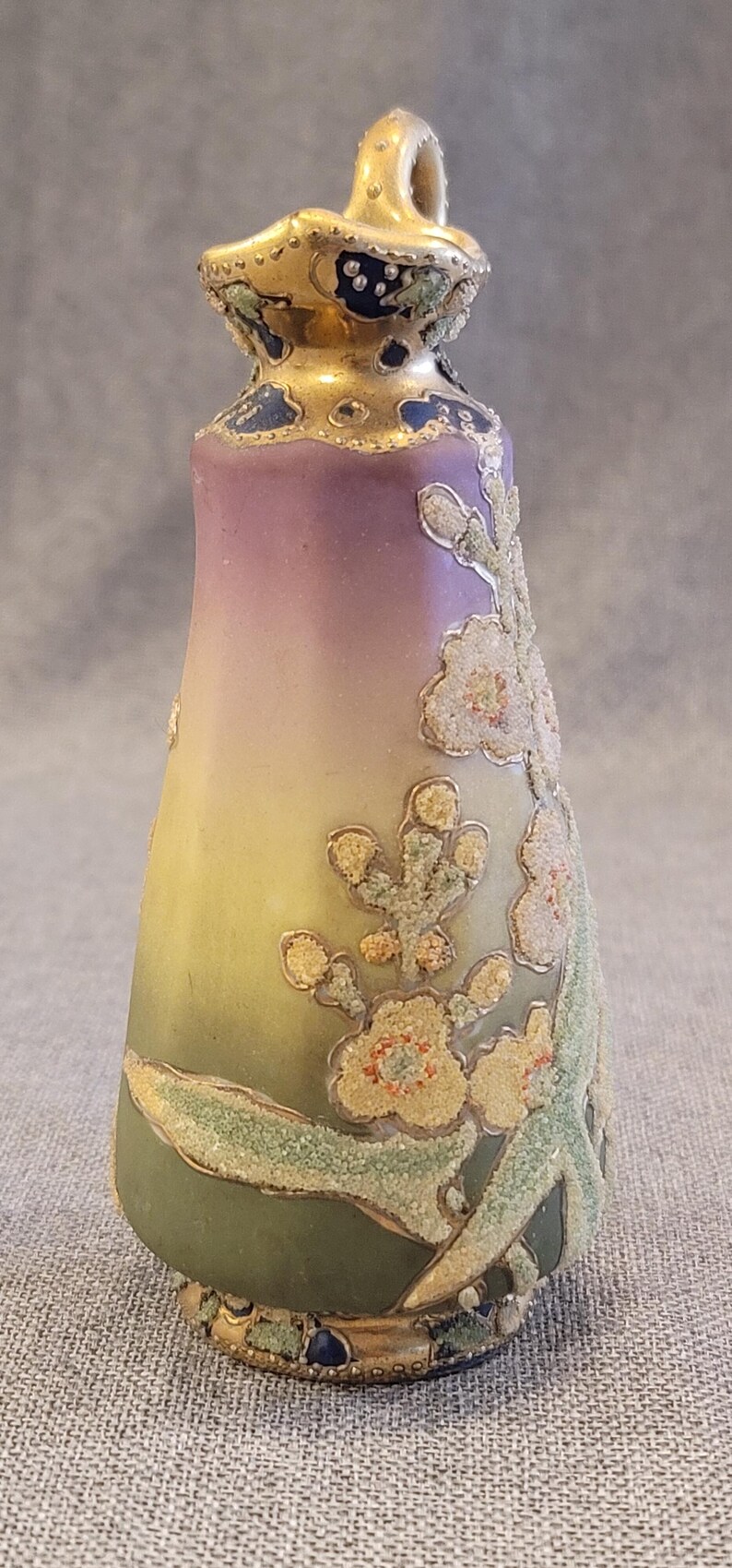 May include: A small, hand-painted ceramic pitcher with a gold handle and floral design. The pitcher has a gradient of colors from purple to yellow to green, with gold accents and white flowers.