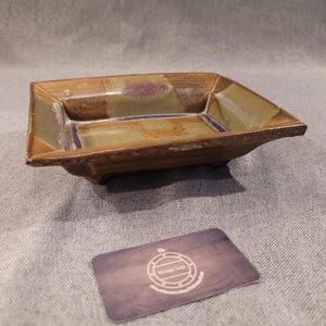 Puede incluir: Un plato cuadrado de cerámica hecho a mano, con exterior marrón e interior verde y morado. El plato tiene una base ligeramente elevada y un borde texturizado. El plato se utiliza probablemente para servir o exhibir.